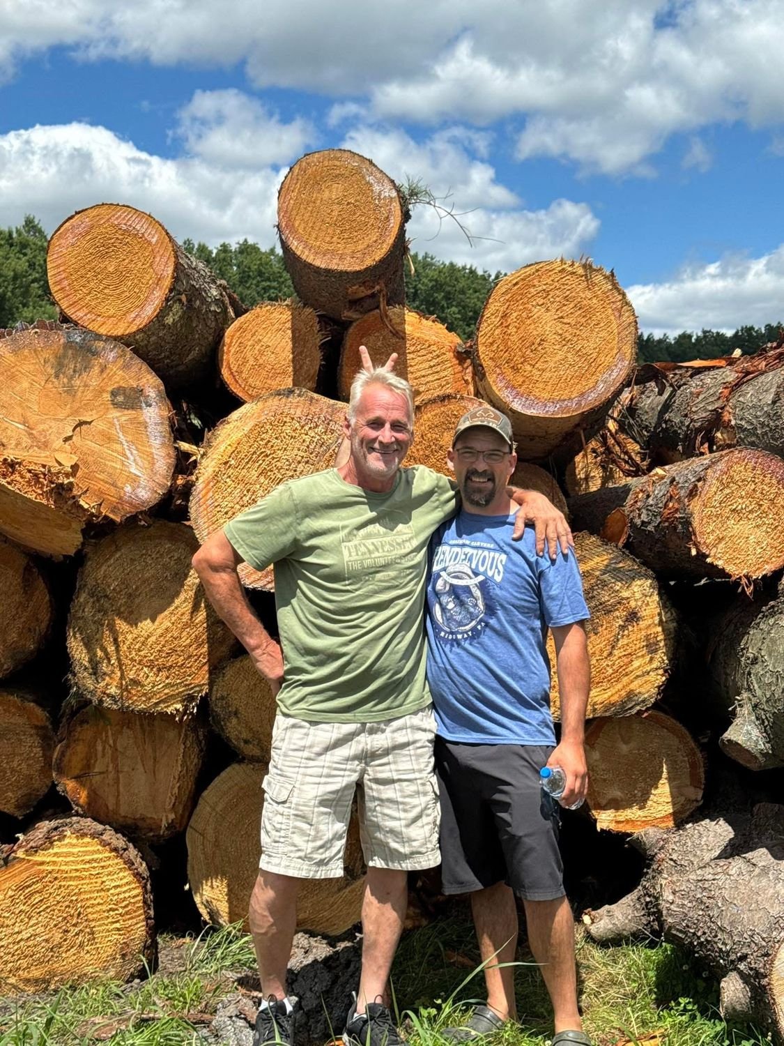 Two men standing in front of a large stack of cut logs.